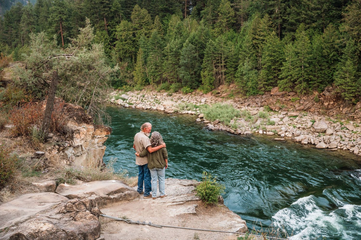 Elderly couple embracing on rocky riverbank surrounded by lush pine forest and turquoise mountain river.