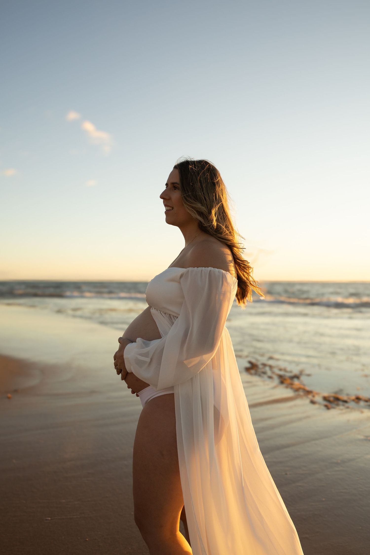 Expectant mother cradles her belly on the shoreline during a golden sunset maternity session at Mandurah Beach.
