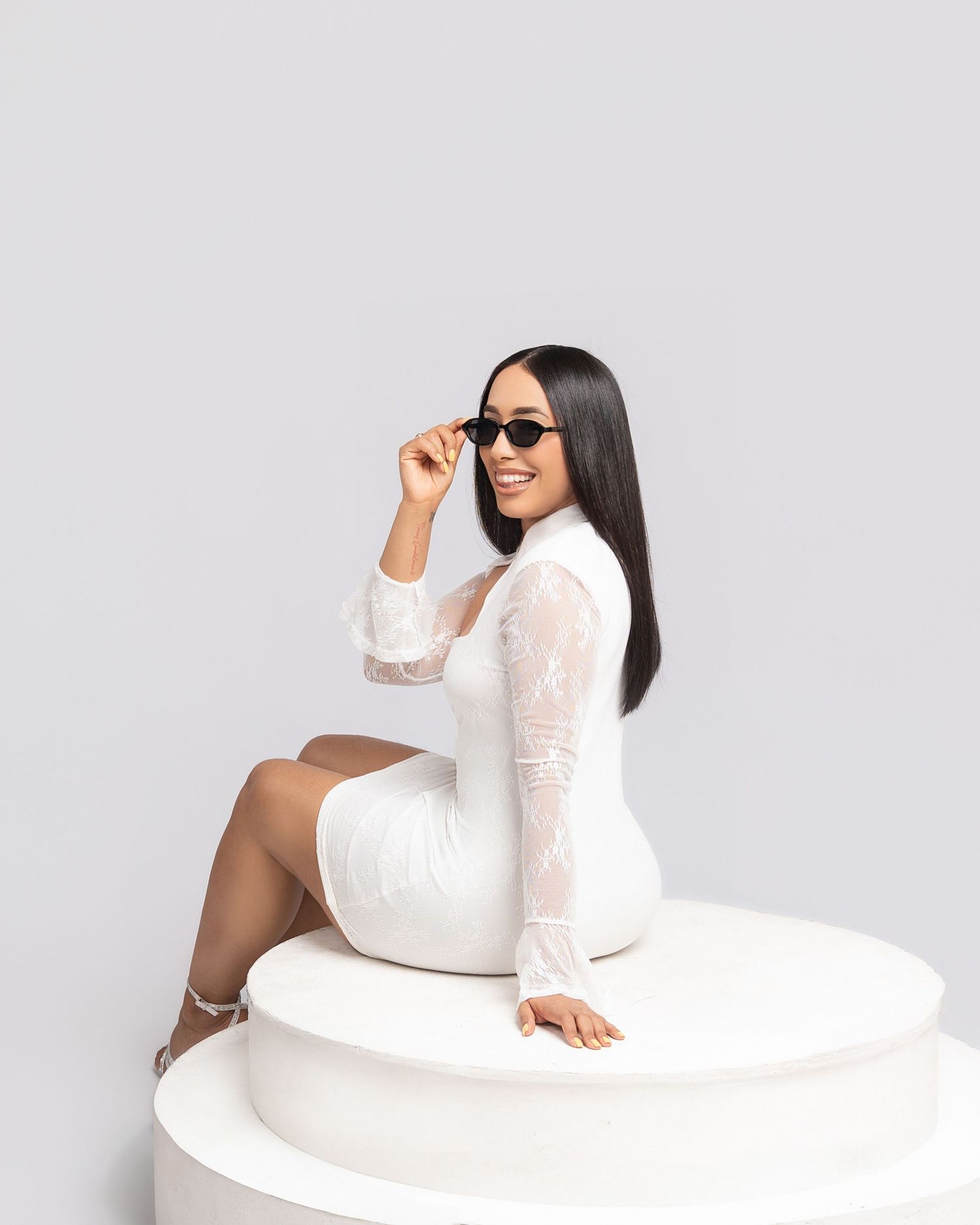 Woman in white lace dress sitting on pedestal, holding black sunglasses, smiling against gray background.