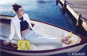 ST&SAT model in white blazer sitting in boat holding yellow handbag with marina background.