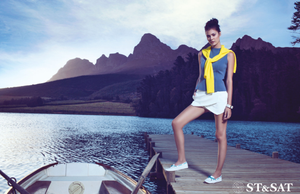 ST&SAT fashion model in yellow top and white skirt posing on dock with mountain lake backdrop.