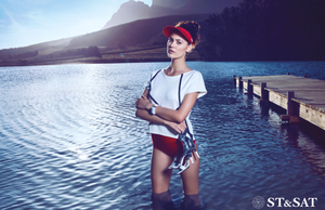 ST&SAT model in red cap and white tee standing in shallow lake water near wooden pier.