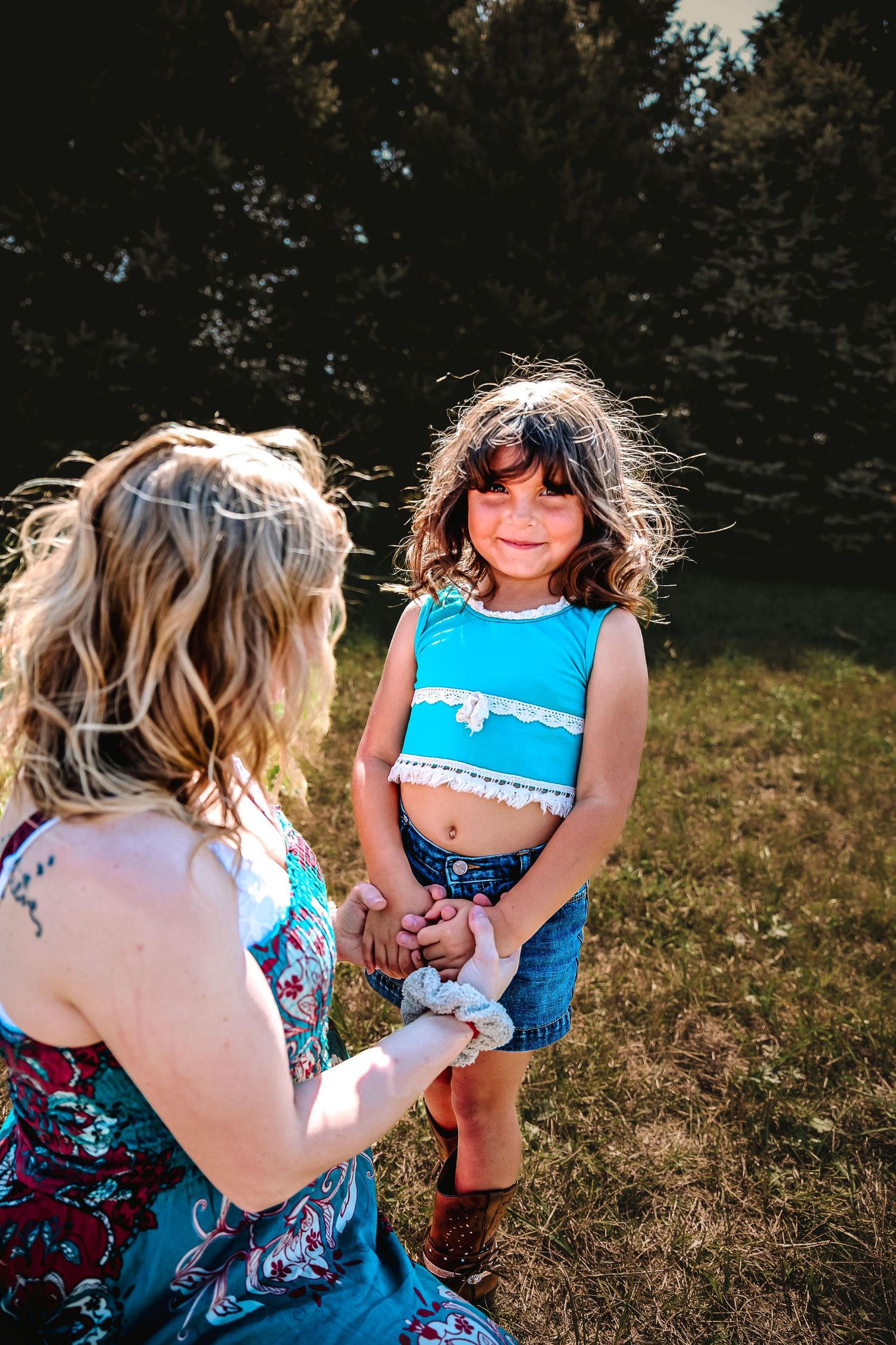 A woman kneels in a sunny field holding hands with a smiling young girl in a blue crop top and denim shorts.