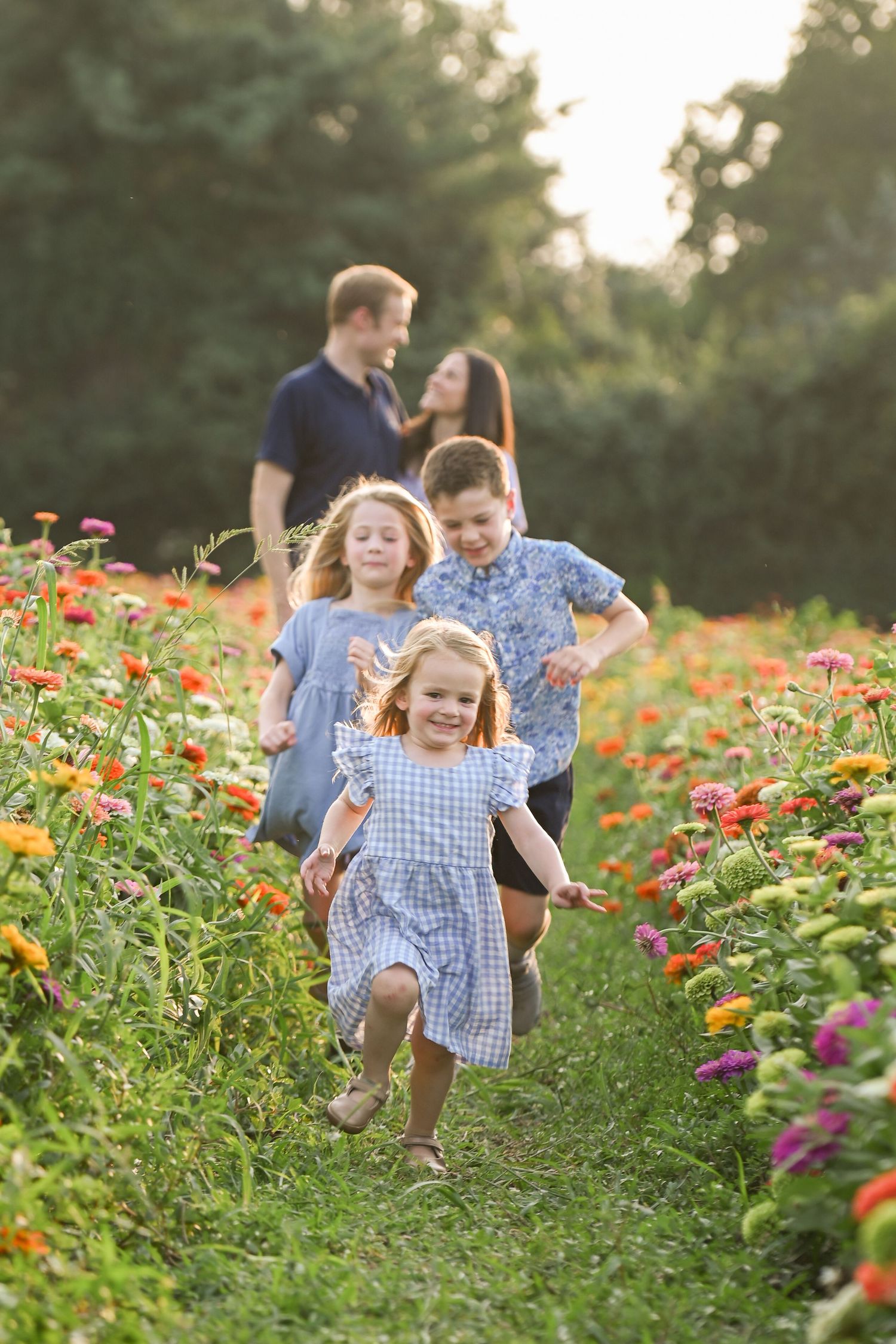 Three children run through a colorful flower field toward the camera as their parents stand together smiling in the background.