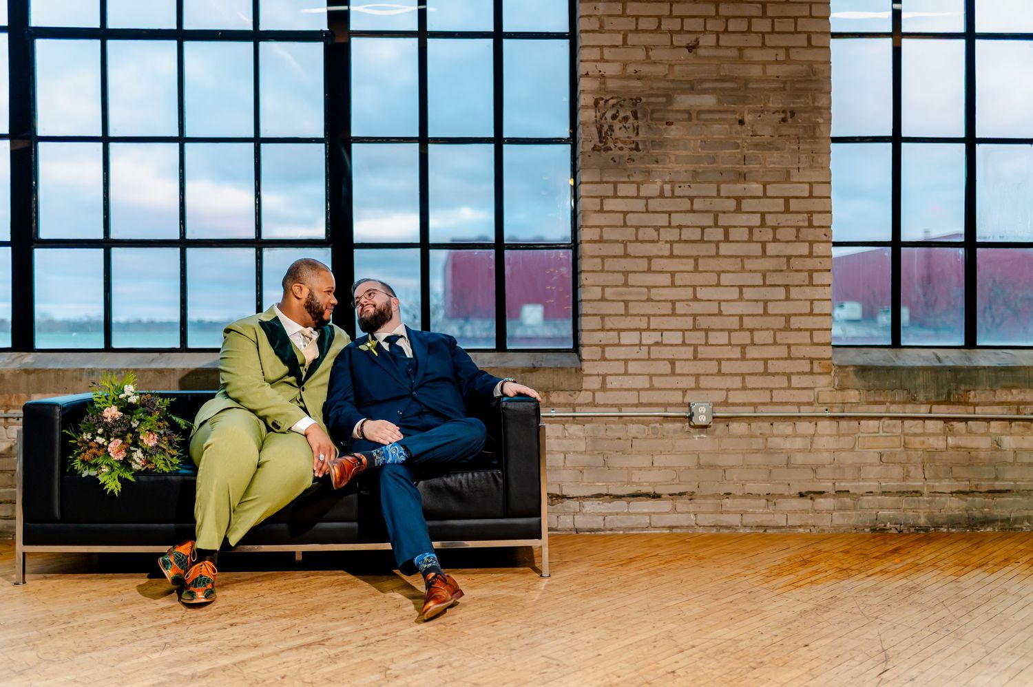 Two grooms in suits sit together on a black couch in an industrial loft venue with large windows.