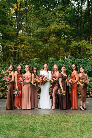 Bride in white gown poses with eight bridesmaids in mismatched earth-tone dresses holding colorful bouquets outdoors.