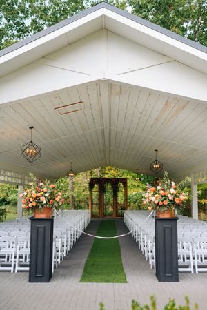 Elegant outdoor wedding chapel with white chairs, green aisle runner, orange floral arrangements, and hanging geometric lanterns.