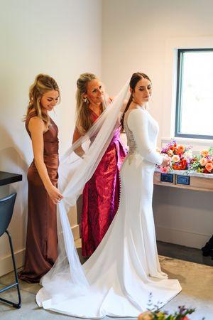 Bride in white gown and veil being assisted by two women in mauve and red dresses before wedding ceremony.