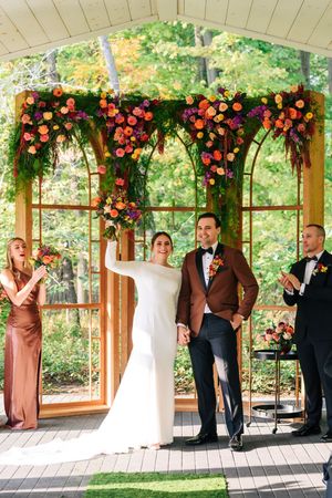 Bride and groom celebrate at outdoor wedding ceremony under floral arch with vibrant orange and purple blooms.