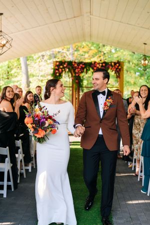 Newlyweds walk down the aisle holding hands, bride in white gown with colorful bouquet, groom in brown tuxedo jacket.