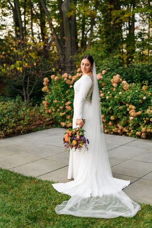 Bride in long-sleeve white gown with cathedral veil holds colorful bouquet in autumn garden setting.