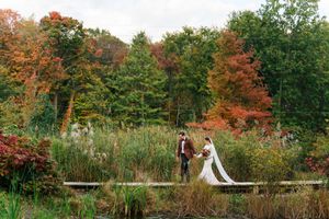 Bride and groom hold hands on a wooden bridge surrounded by vibrant autumn foliage in a lush garden setting.