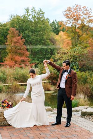 Bride and groom dancing outdoors in autumn garden, she in white gown, he in brown tuxedo jacket.