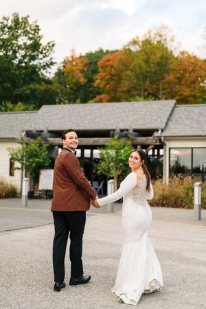 Bride in white long-sleeve gown and groom in brown tuxedo hold hands outdoors during fall wedding portrait.