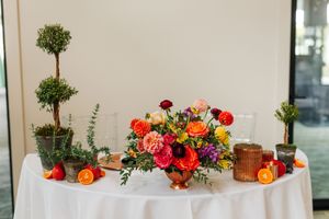 Colorful wedding centerpiece with orange, red, and purple flowers, topiaries, and halved oranges on a white tablecloth.