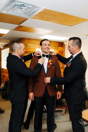 Groom in brown tuxedo jacket smiles as two groomsmen in black suits adjust his bow tie and boutonniere.