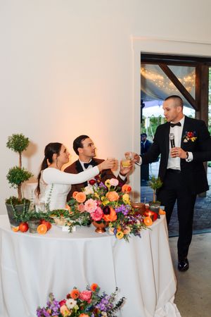Bride and groom toast with a guest at reception table adorned with colorful floral arrangements and citrus decor.