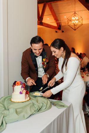 Smiling bride and groom in elegant attire cut their decorated white wedding cake at indoor reception.