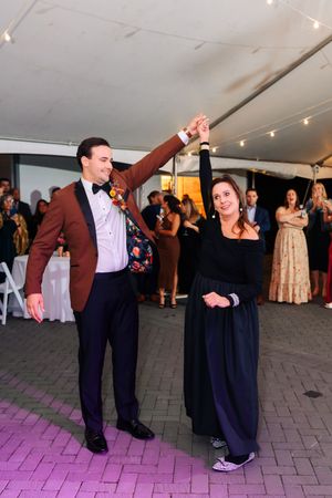 Groom in burgundy tuxedo jacket twirls woman in black gown on wedding reception dance floor under tent lights.