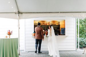 Bride and groom at outdoor wedding bar under white tent, ordering drinks at rustic white shiplap bar.