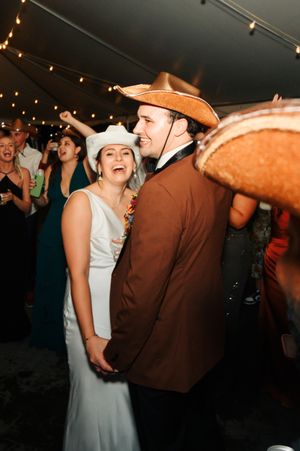 Bride and groom in cowboy hats dancing joyfully at wedding reception under string lights.