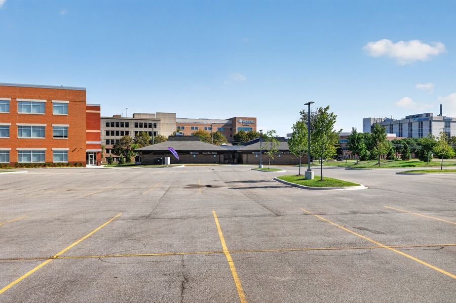 Empty parking lot with yellow lines near brick buildings and trees under a clear blue sky.