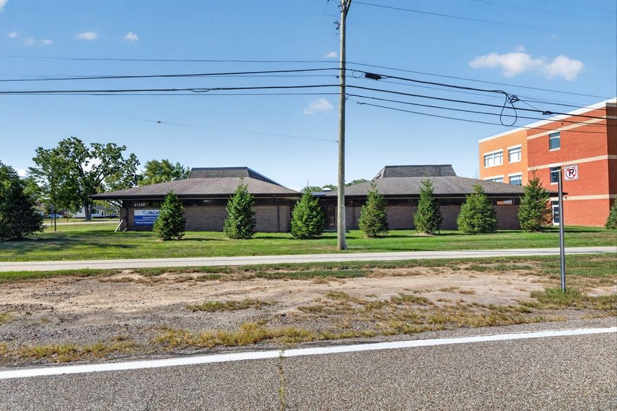 Single-story commercial building with dark roof panels and brick facade, situated along a suburban road with green trees.