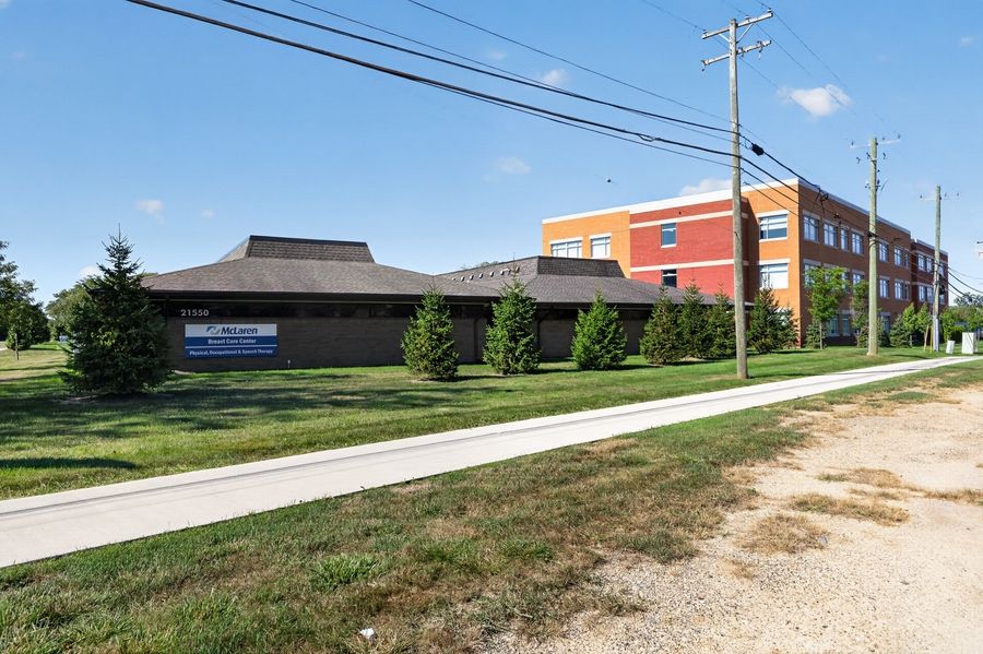 Low-rise commercial building with brick facade beside a multi-story red brick structure, surrounded by green lawns and evergreen shrubs.