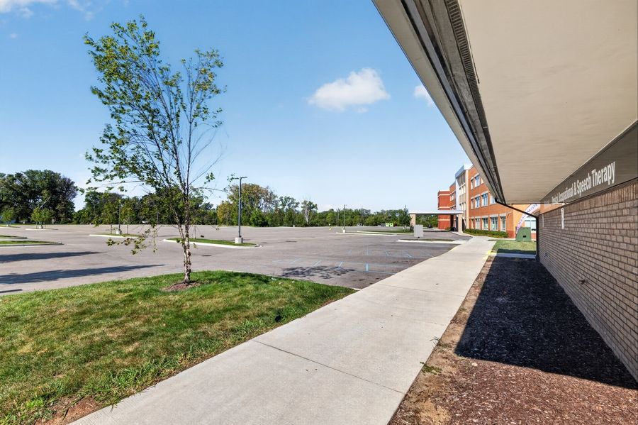 Empty school parking lot with newly planted trees, sidewalk, and red brick building under clear blue sky.