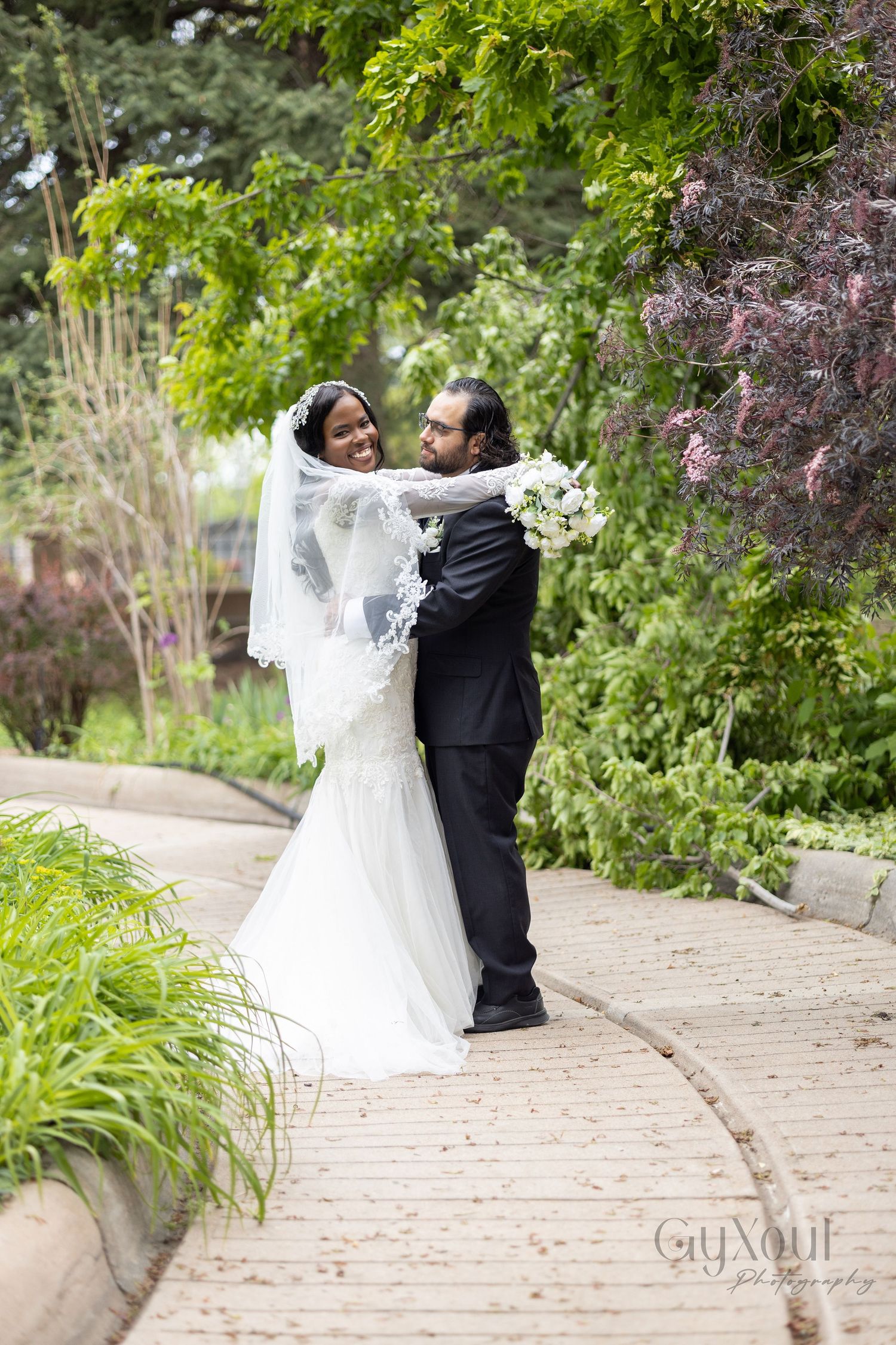 A groom in a black suit lifts his bride in a white gown and veil on a garden path surrounded by lush greenery.