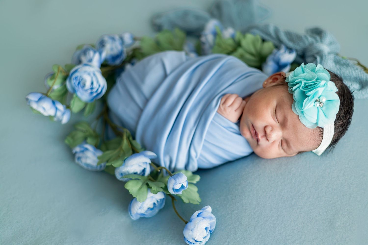 Newborn baby girl sleeping peacefully, swaddled in blue wrap with floral headband, surrounded by blue roses.