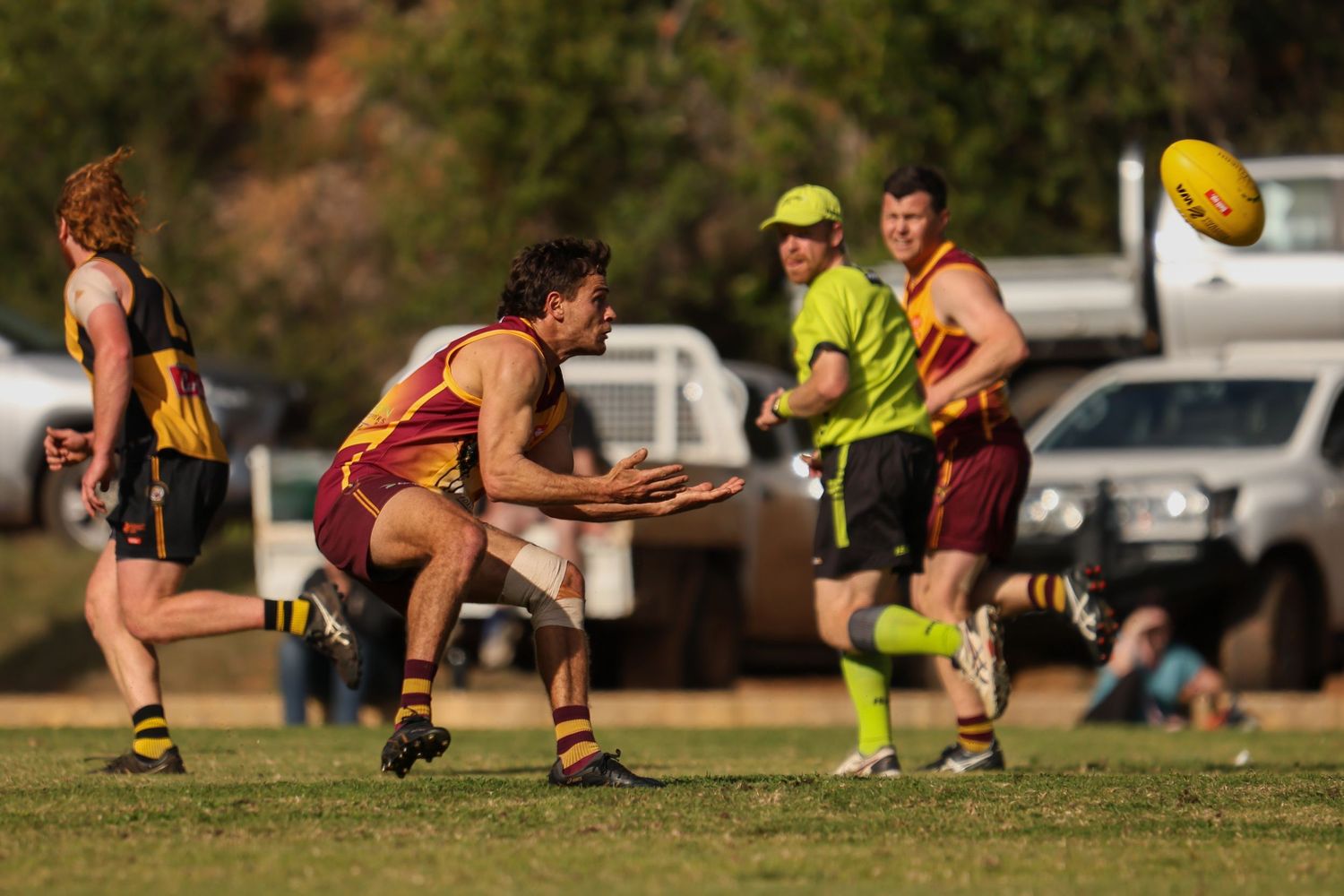 Australian rules football players compete in an outdoor match, with a referee in yellow and a ball visible mid-air.
