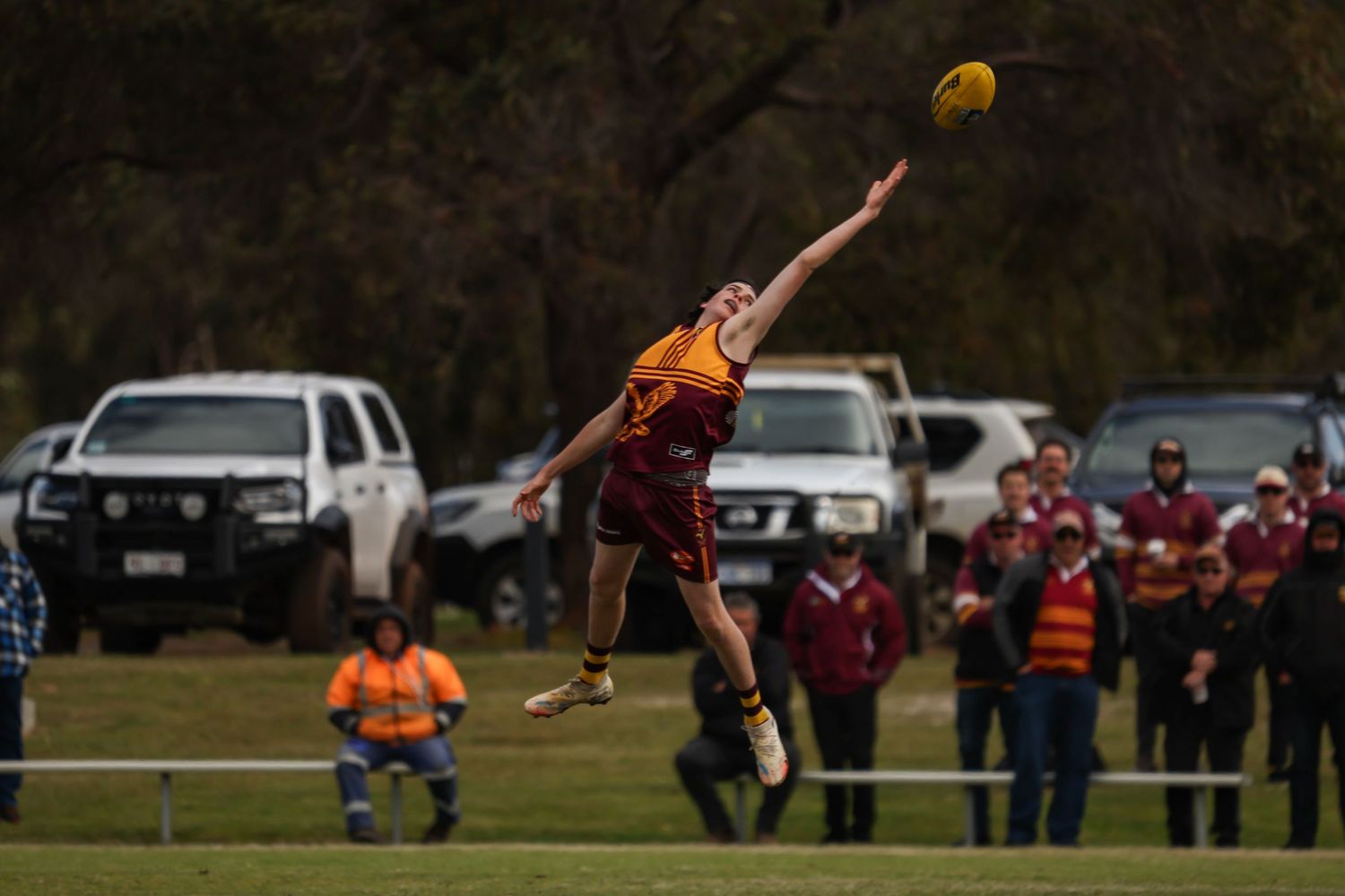 Player leaping high to catch an AFL football during an outdoor match, with spectators and parked cars in the background.