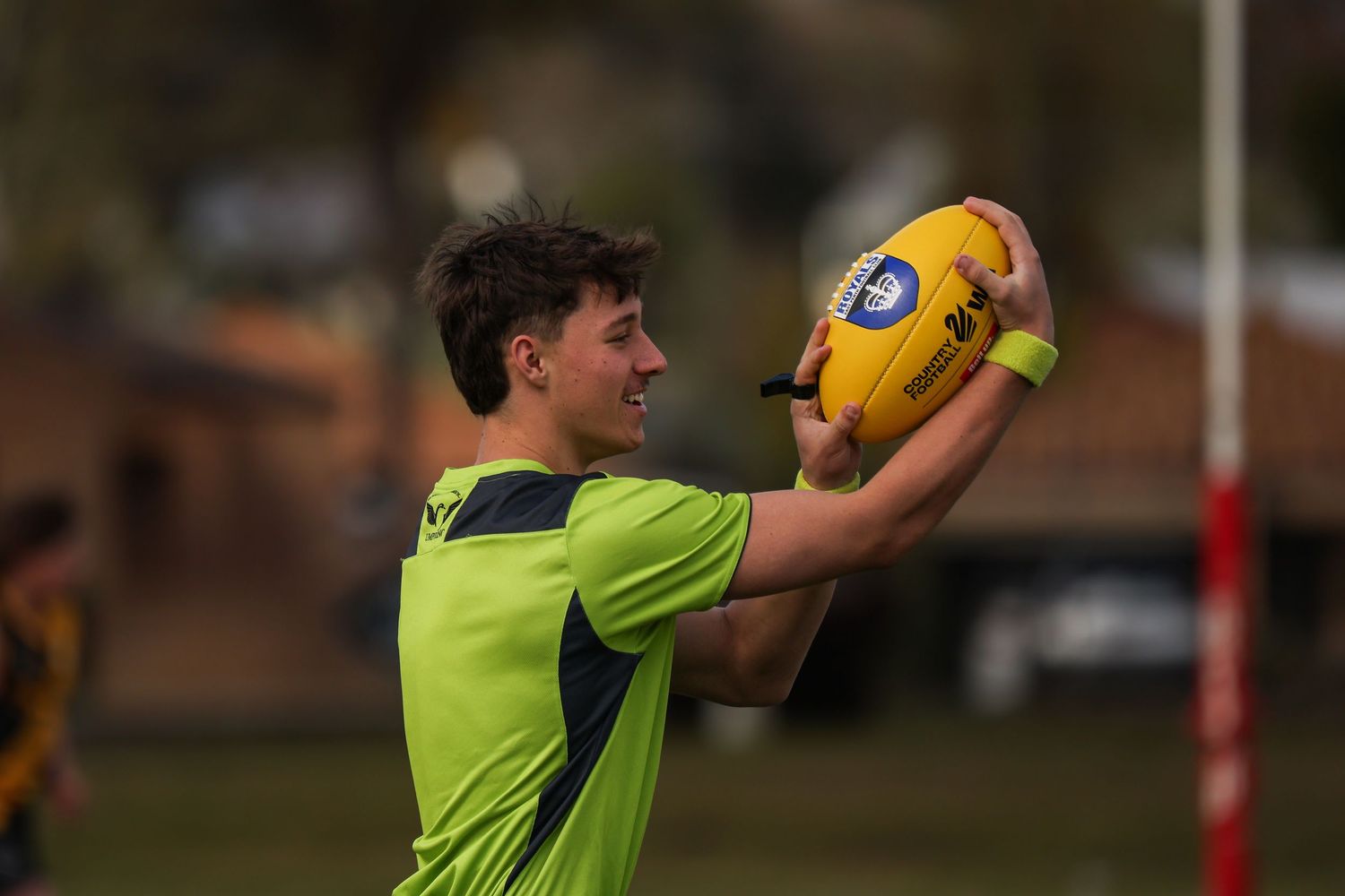 Young male athlete in green jersey holds yellow AFL football above his head on an outdoor sports field.