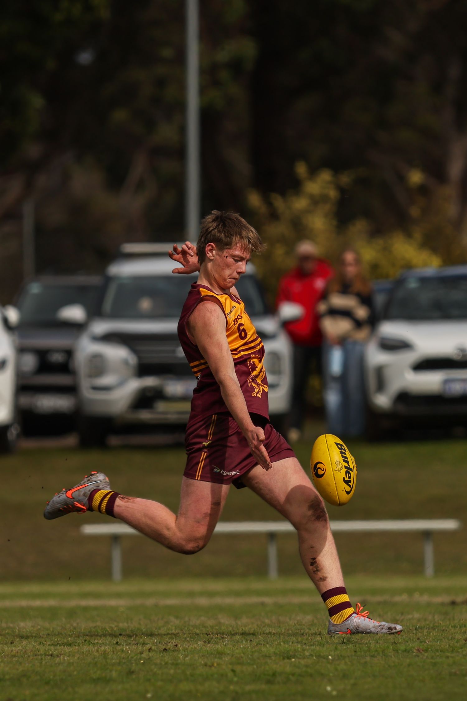 Young AFL player in maroon and gold uniform kicking a yellow football on a green oval during a match.