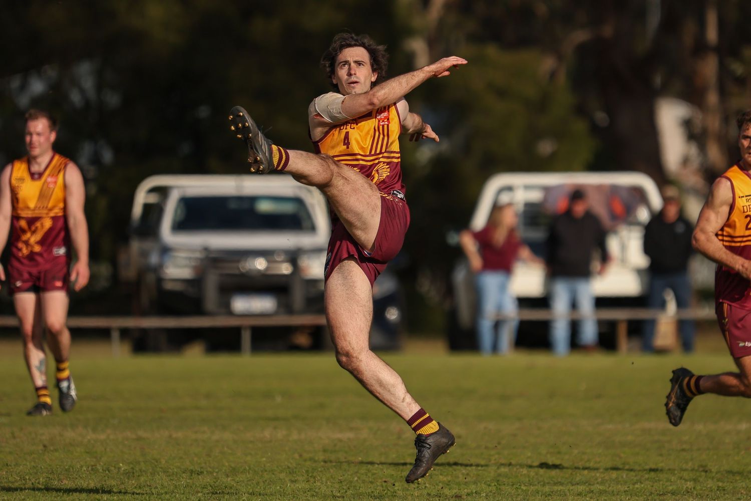 Australian rules footballer in maroon and gold uniform kicking ball mid-air during an outdoor match.