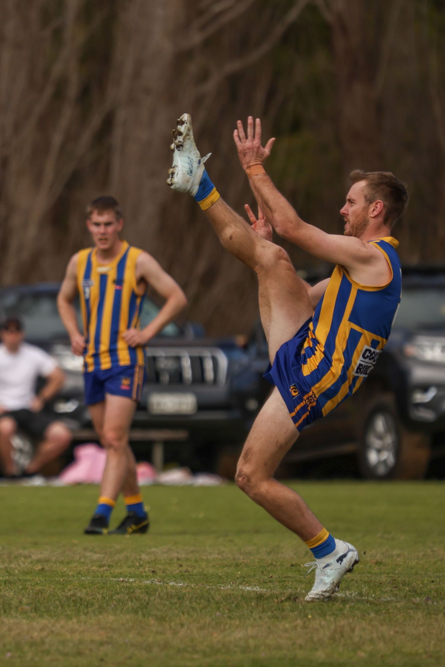 Australian rules footballer in blue and gold uniform kicking ball during outdoor match, teammate visible in background.