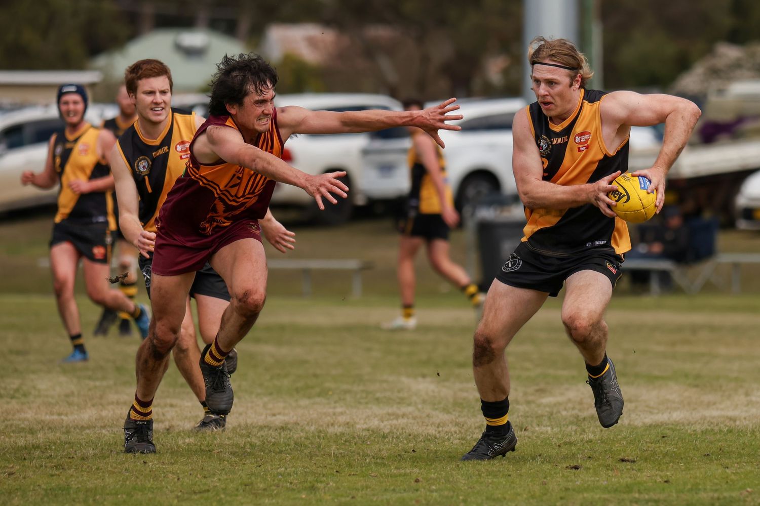 Australian rules football players compete on a grass oval, with one player carrying the ball while evading an opponent.
