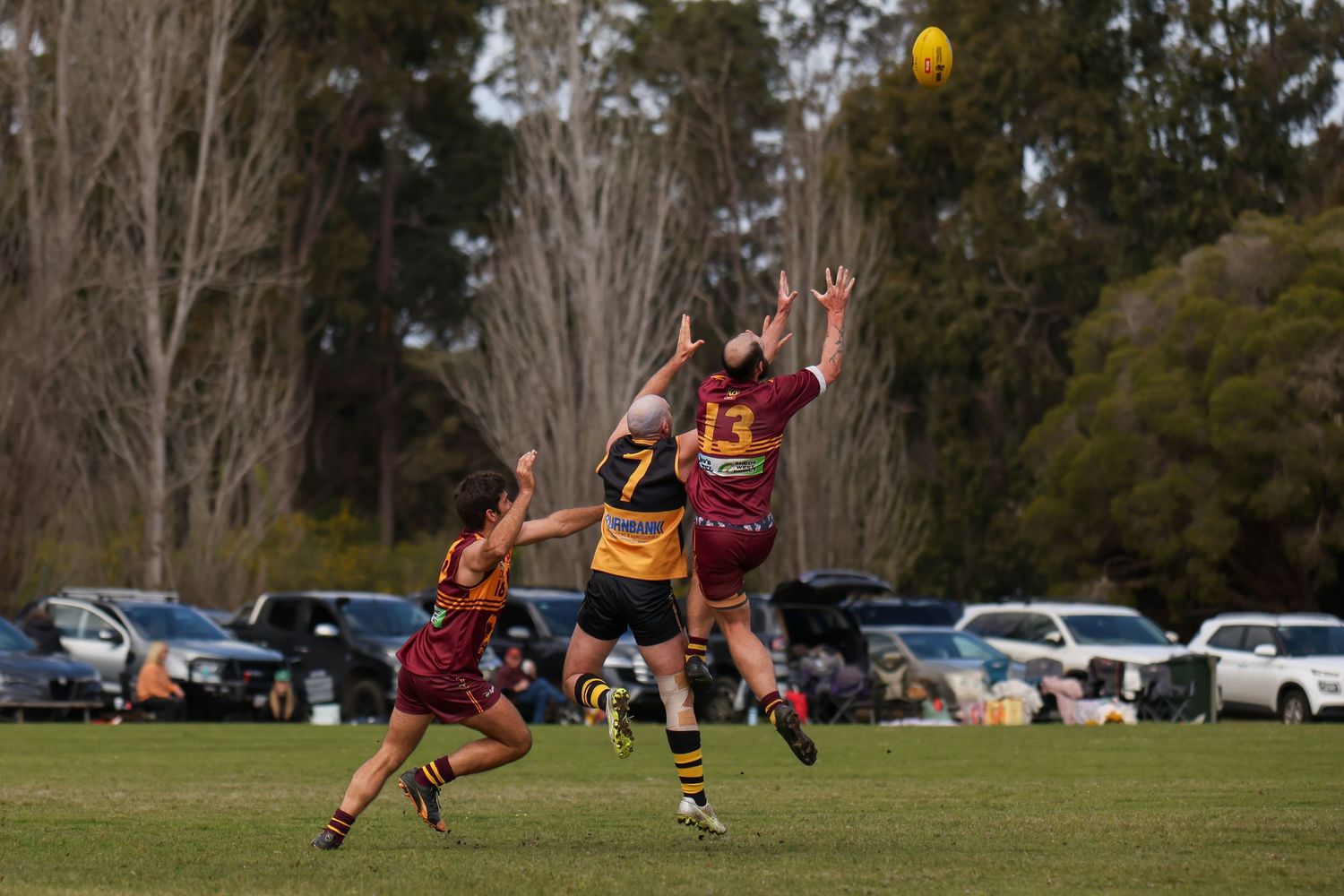 Australian rules football players compete for a mark during an outdoor amateur match on a green oval field.