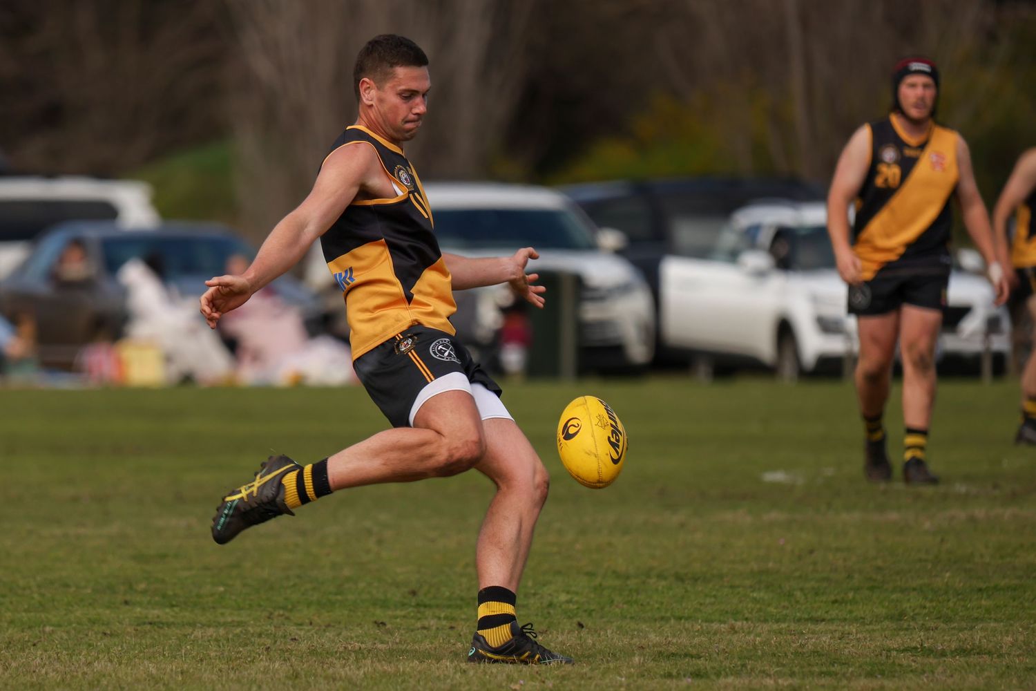 AFL player in yellow and black uniform kicking football during a competitive outdoor match on a green grass field.