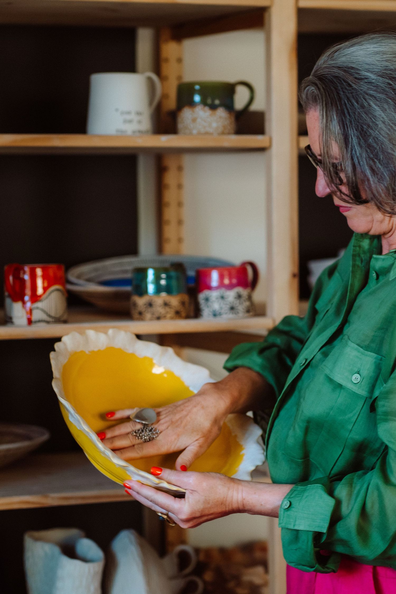 Woman in green shirt examines yellow ceramic bowl in pottery shop with colorful mugs on wooden shelves.