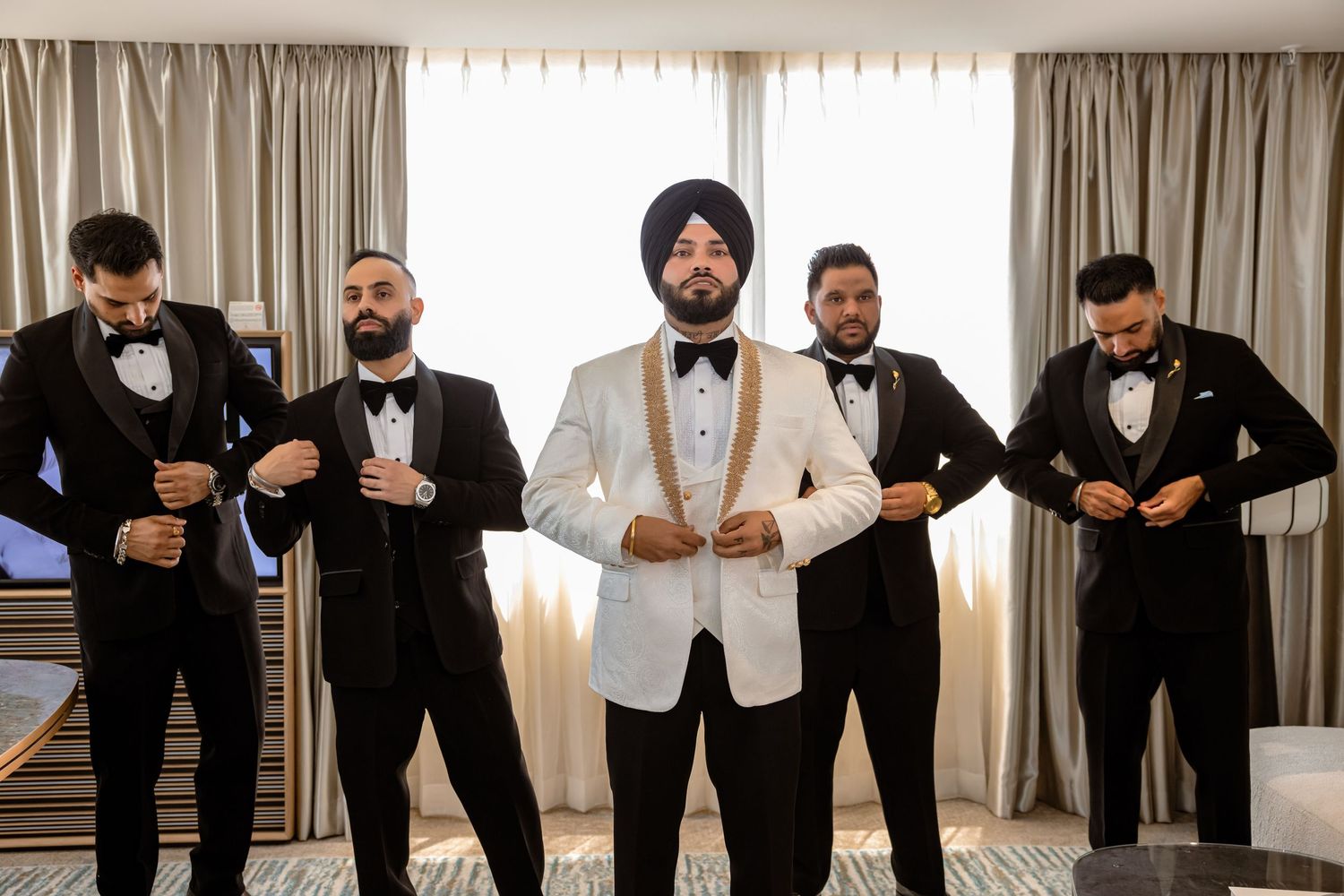 Sikh groom in white tuxedo and turban stands with four groomsmen in black suits adjusting bowties in hotel room.
