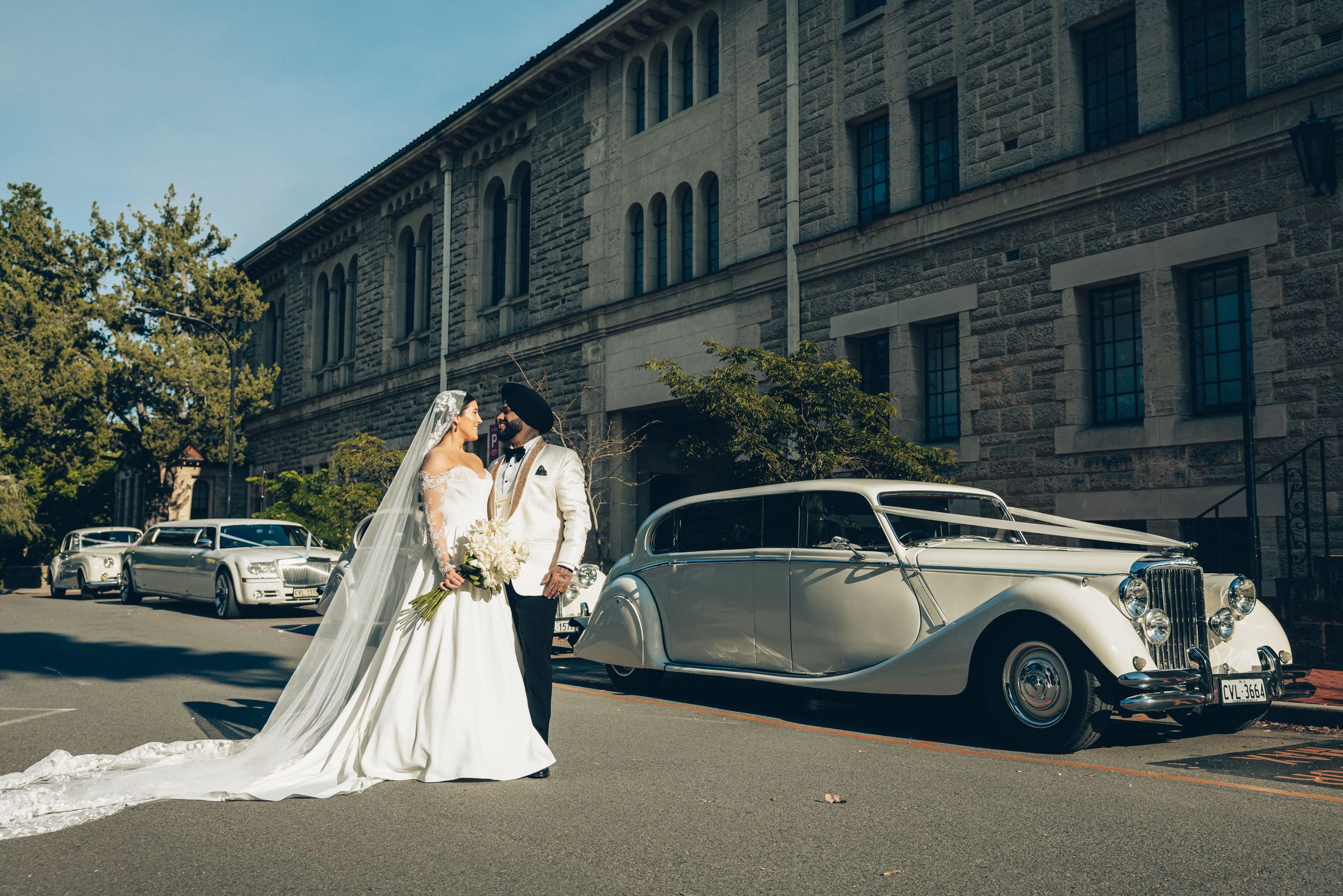 Bride and groom kissing beside a vintage white wedding car on a sunny day outside a historic stone building.