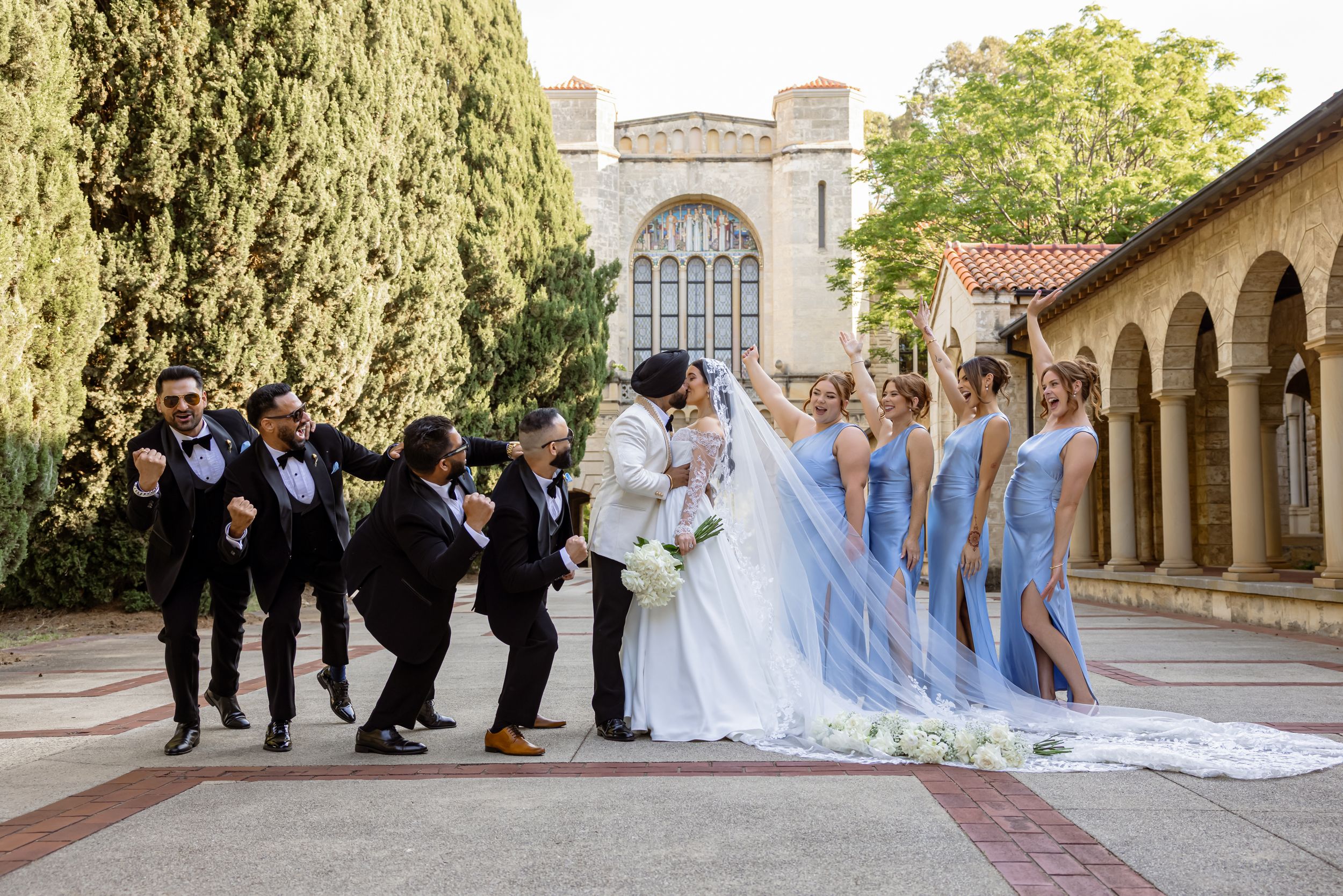Wedding party poses outside a church, groomsmen in tuxedos cheering as bridesmaids in blue gowns flank the bride and groom.