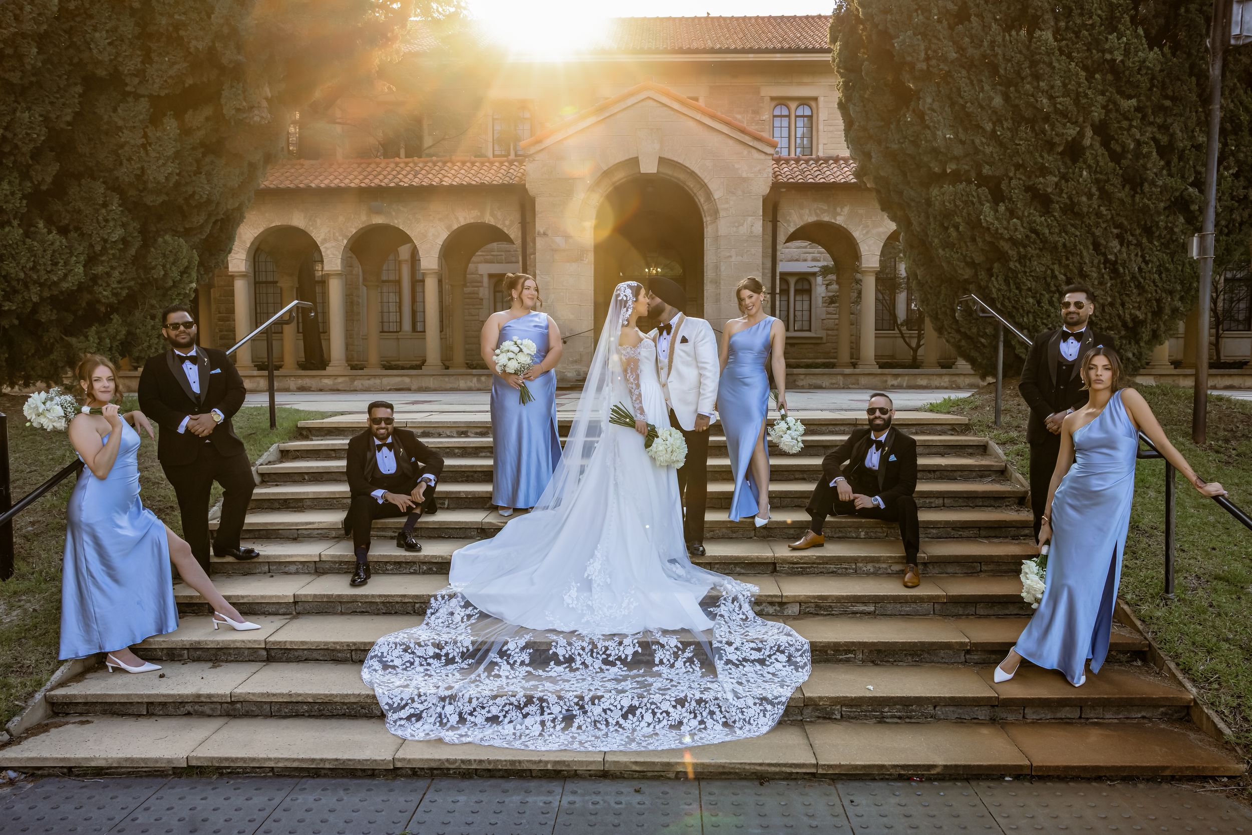 Bride and groom pose with wedding party in blue dresses and black suits on sunlit steps of elegant venue.