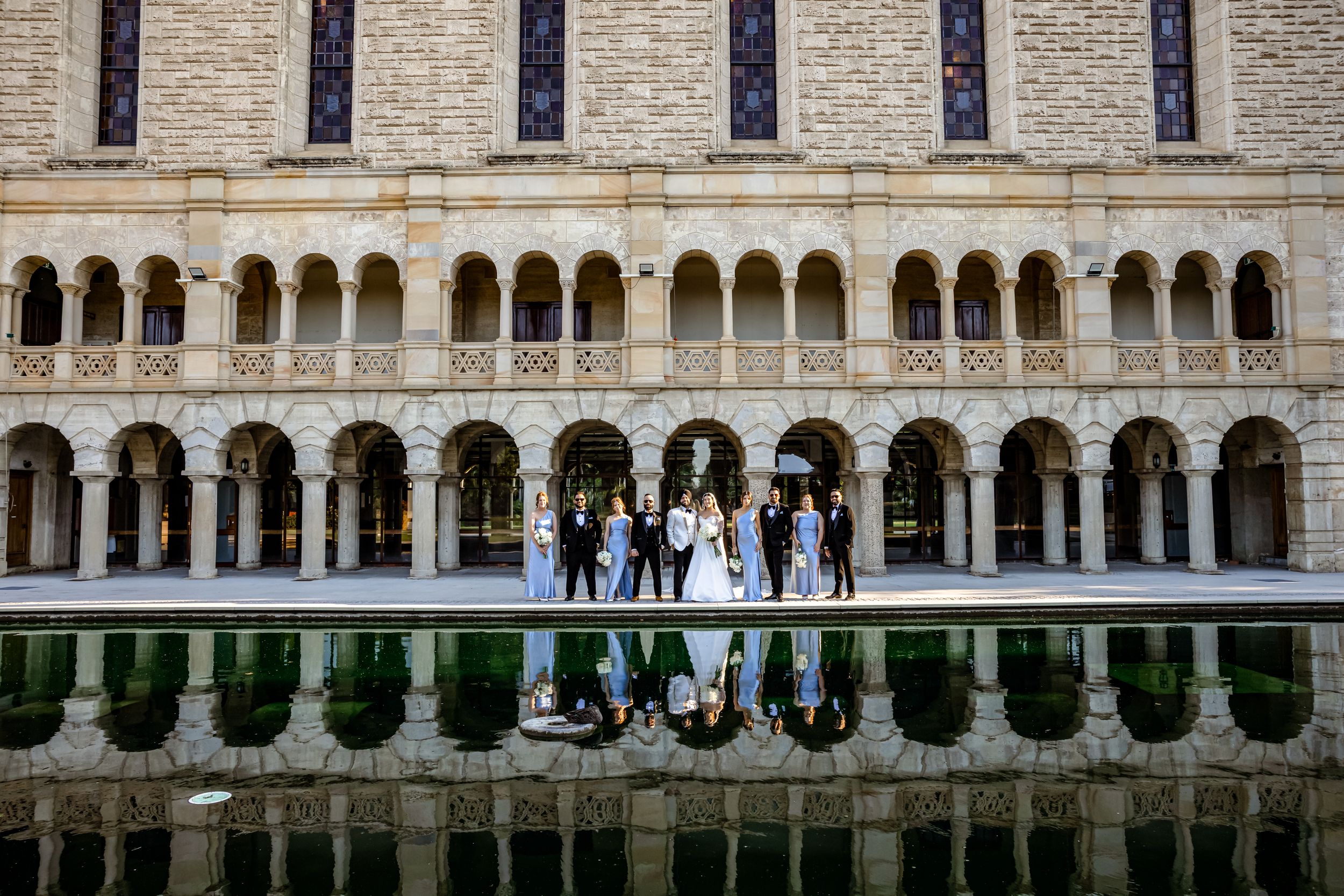 Wedding party poses before ornate arched colonnade building, reflected in still water below.