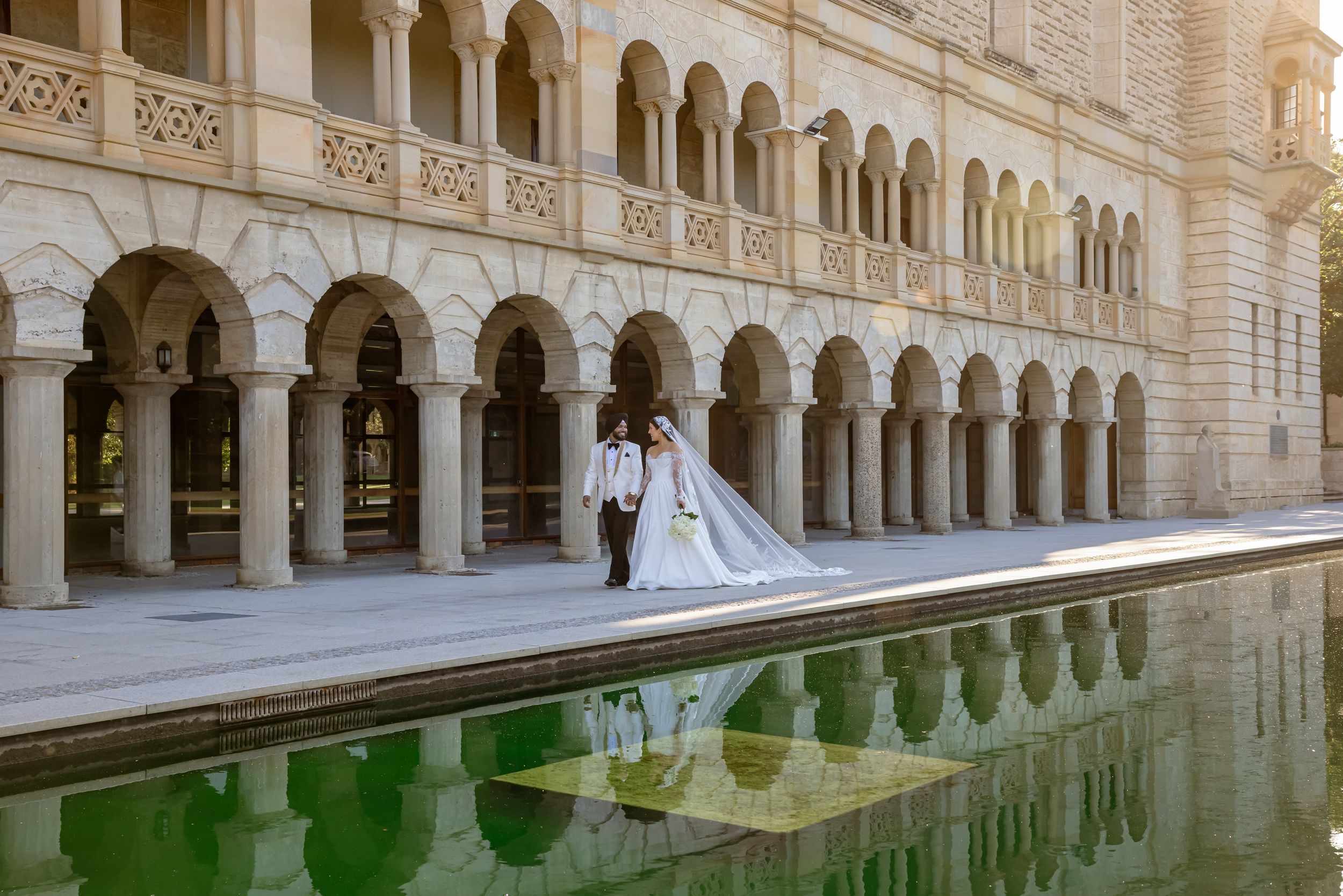 Bride and groom pose by a reflective pool at an ornate arched building in an elegant wedding photo.
