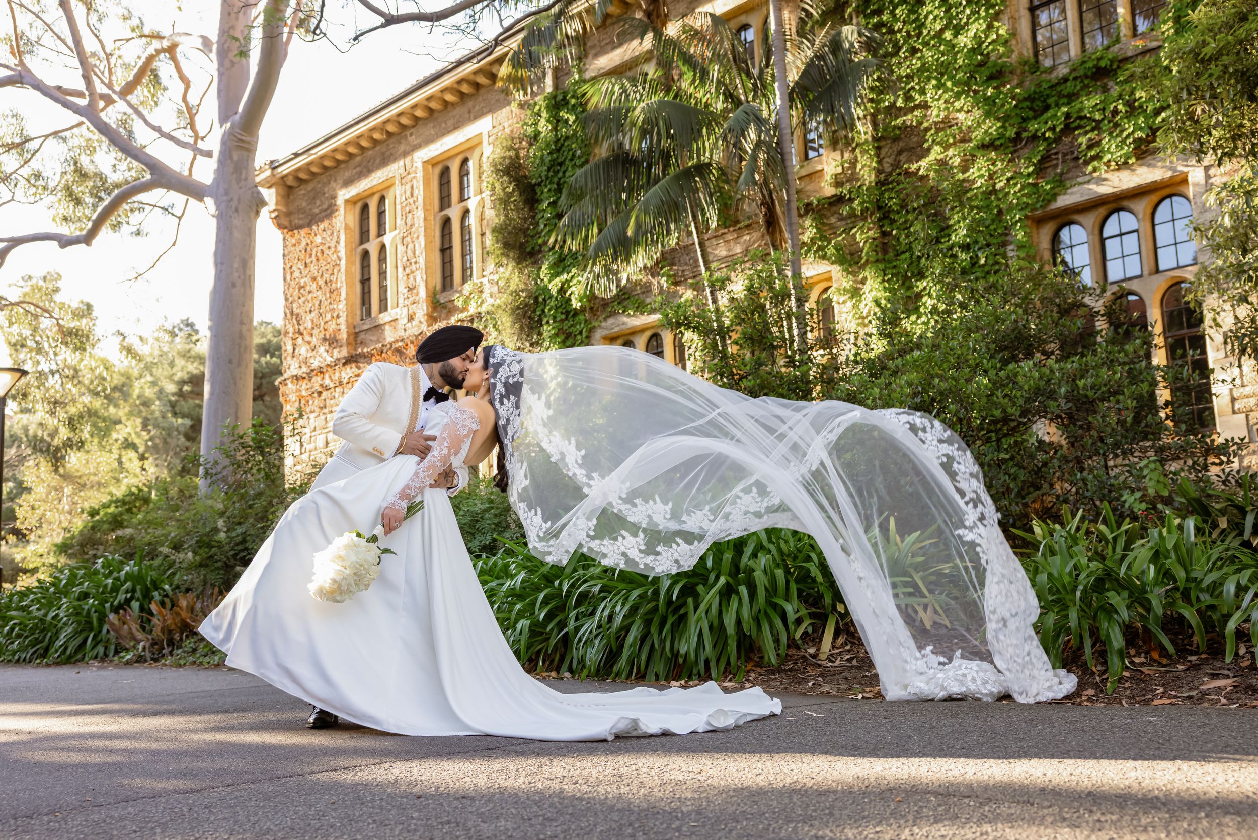Bride and groom share a romantic dip kiss outdoors, with flowing lace veil billowing in the wind at an elegant venue.