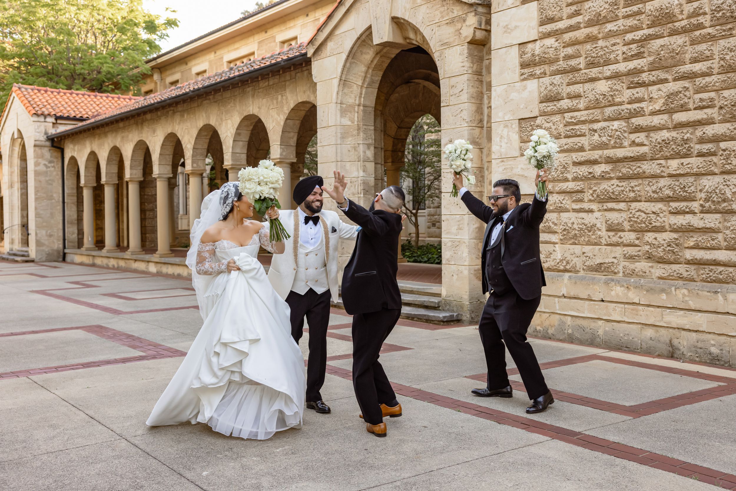 Joyful wedding party celebrating outdoors near stone arched building, bride in white gown holding bouquet with groom and guests.