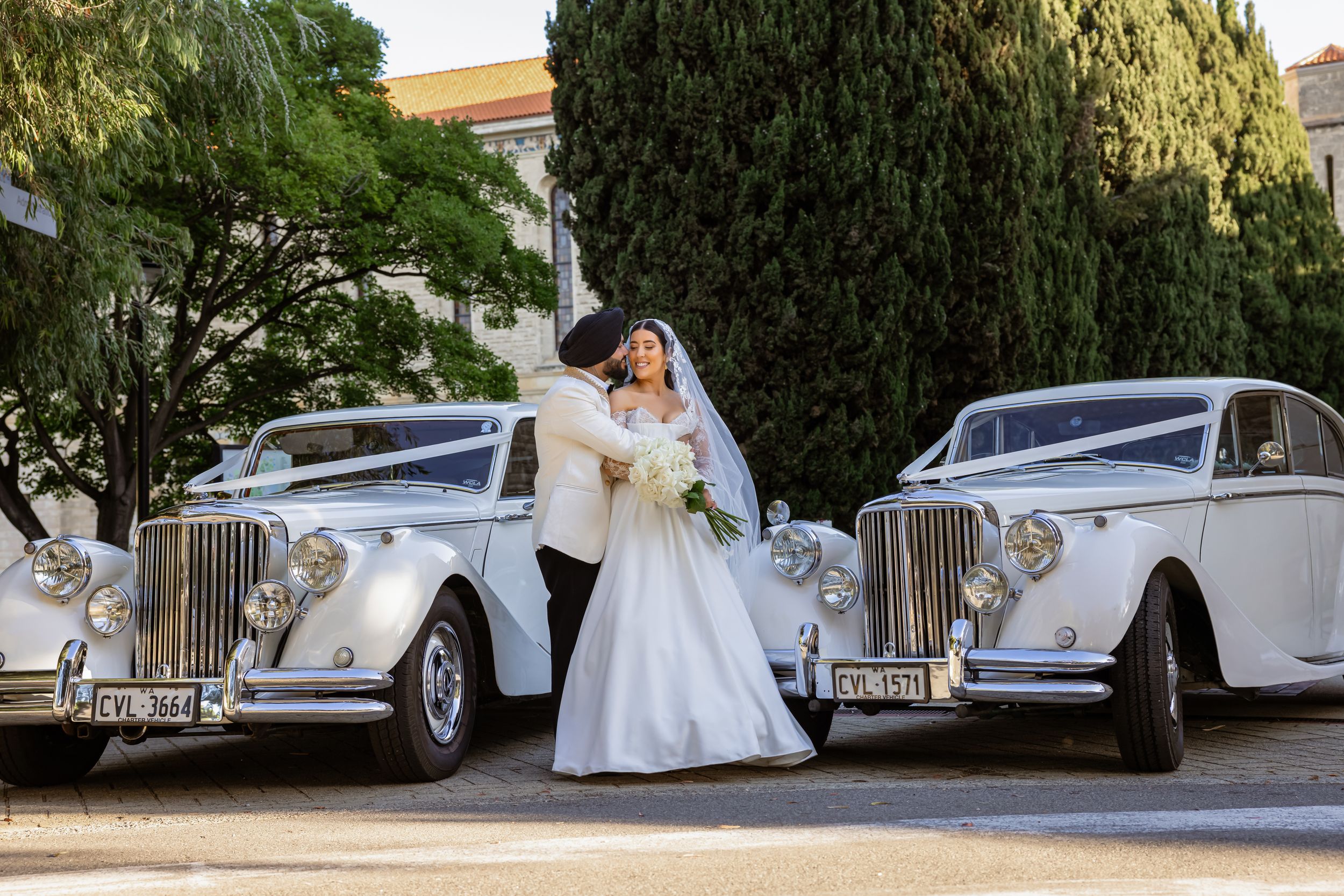 Bride and groom kissing between two classic white vintage wedding cars decorated with white ribbons outdoors.